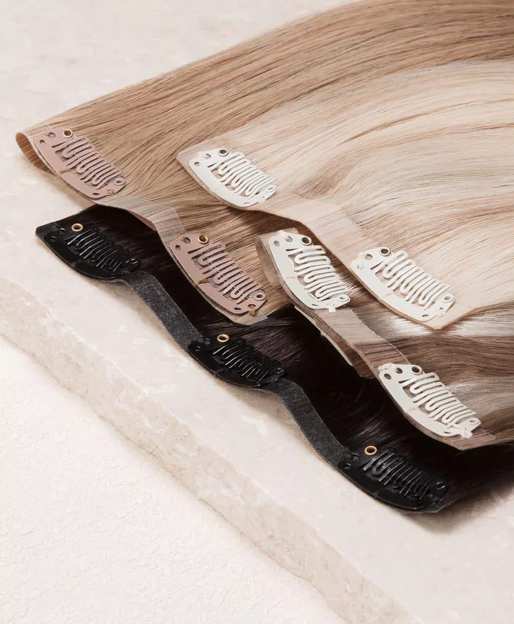 Drei Clip-in-Extensions in Blond, Aschbraun und Schwarz mit farblich passenden Clips auf heller Steinplatte