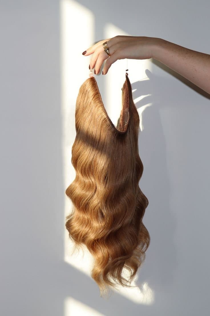 A hand holding a halo hair extension by its wire, with long golden-brown waves hanging down