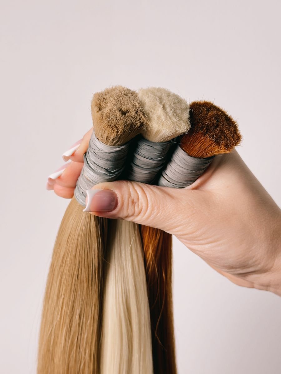 A hand holding three bundles of extension hair in light brown, blonde and copper