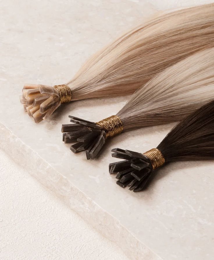 Flat Tip: jede Strähne endet in einer Keratin-Spitze Drei Flat-Tip-Extensions in Blond und Braun mit Keratin-Spitze und goldener Bindung auf heller Steinplatte