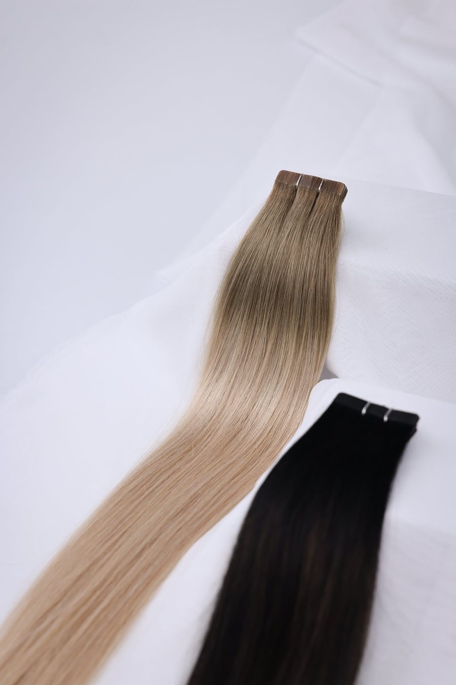 Tape-in in Aschblond und Schwarz Zwei Tape-in-Extensions, eine aschblond und eine schwarz, auf weißem Stoff